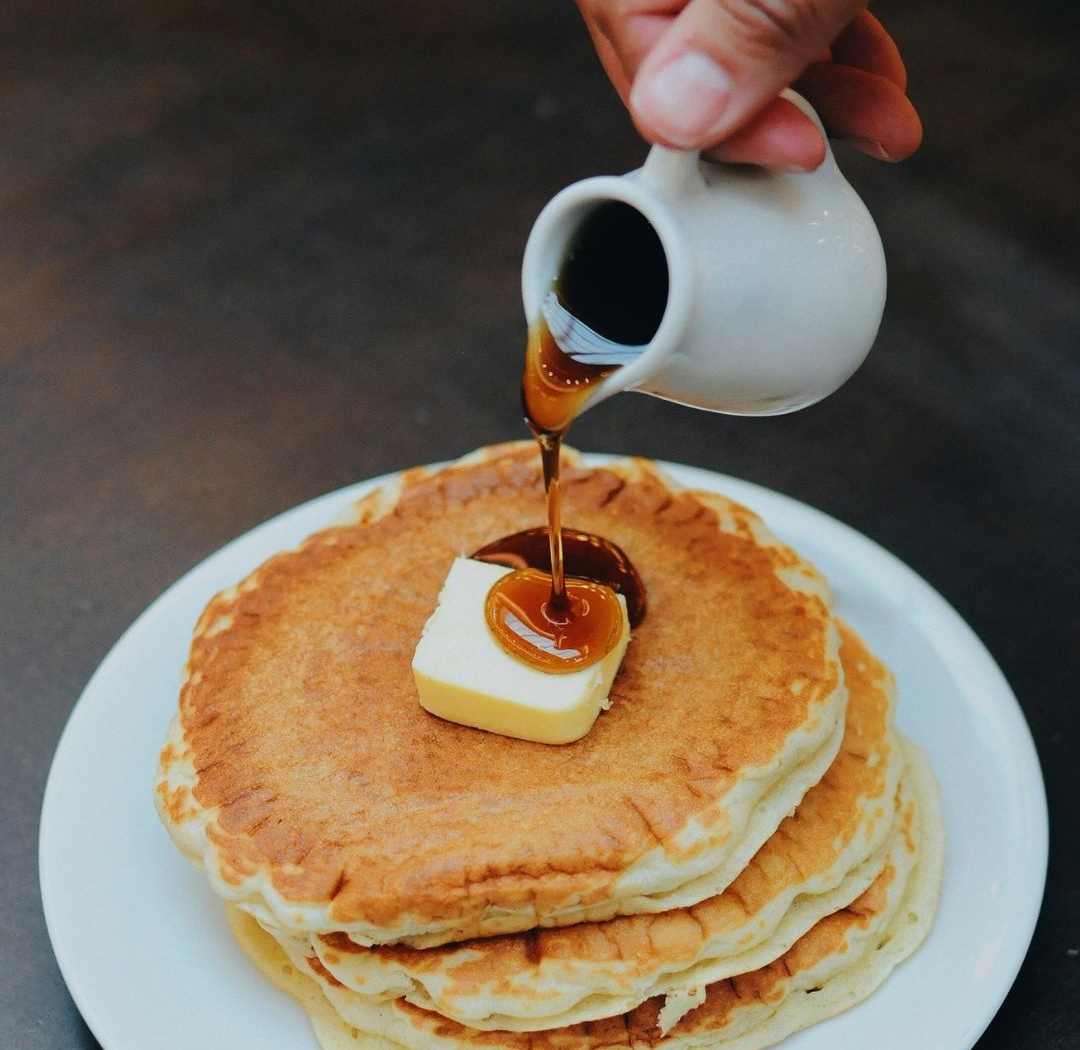 Syrup being poured over a stack of pancakes on a white plate.