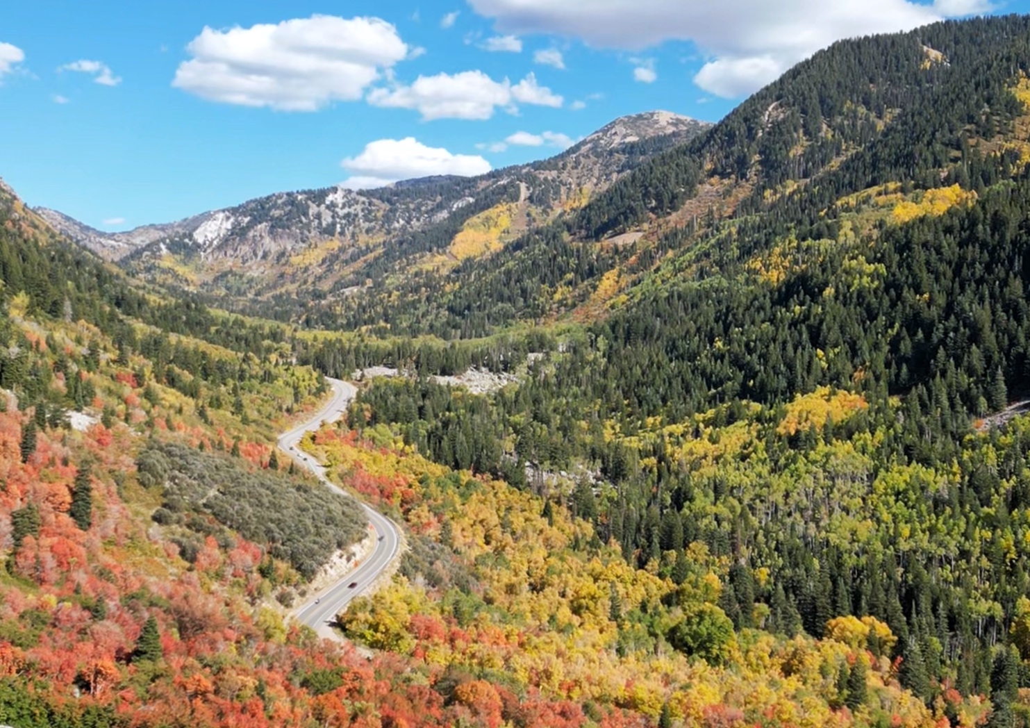 Winding mountain road snaking through a valley filled with vibrant red, orange, and yellow autumn foliage under a blue sky.