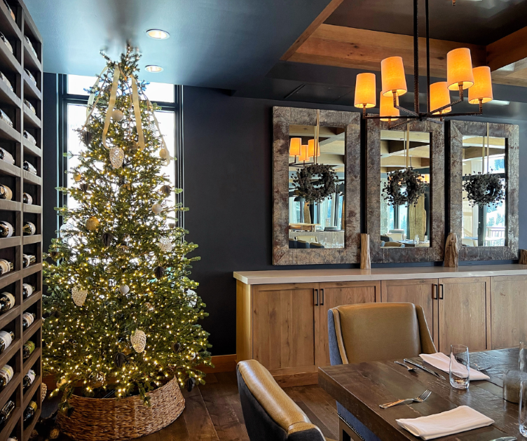 Interior of an elegant dining room decorated for the holidays, featuring a tall lit Christmas tree next to a wine rack, and three rustic mirrors adorned with wreaths hanging above a sideboard.