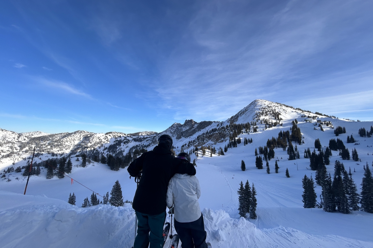 Two people standing on a snowy ridge admiring the mountain view.