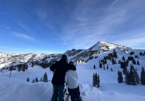 Two people standing on a snowy ridge admiring the mountain view.