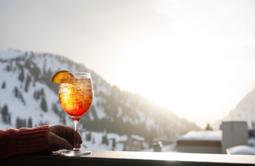 Hand holding an orange spritz cocktail against a snowy mountain background.