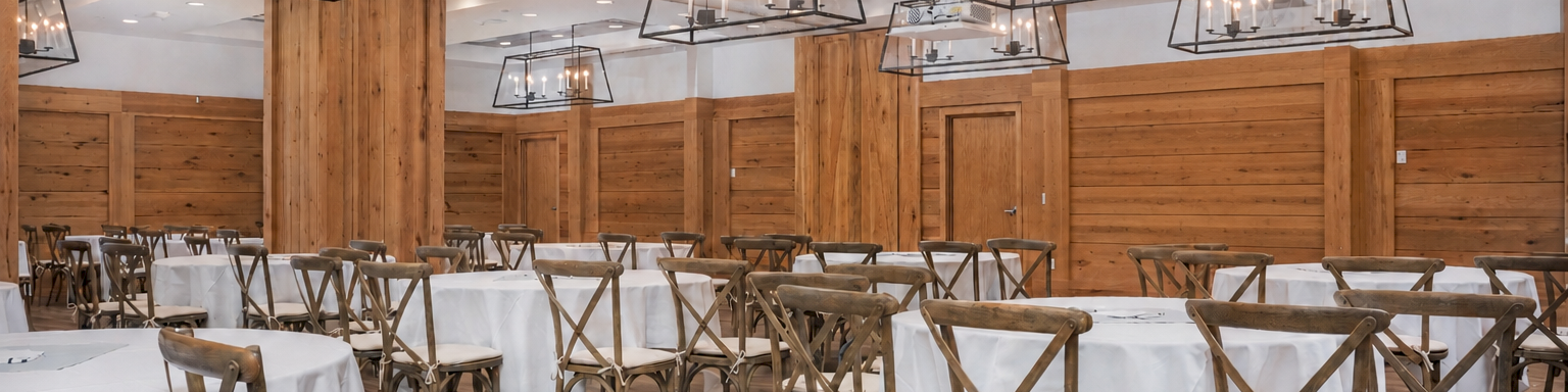A banquet hall set with white tablecloths, wooden X-back chairs, and hanging lantern-style chandeliers in a wood-paneled room.