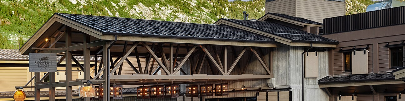 A mountain lodge with timber beams, stone base, and a dark metal roof under clear skies.