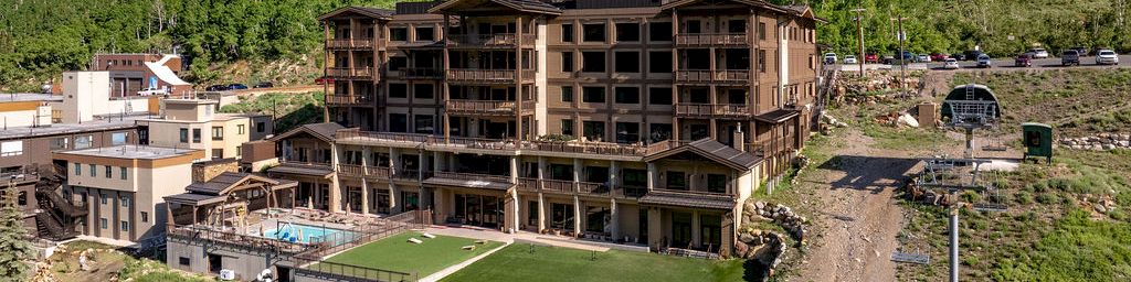 Mountain resort buildings with a central grassy lawn and balconies, surrounded by pine-covered hills.