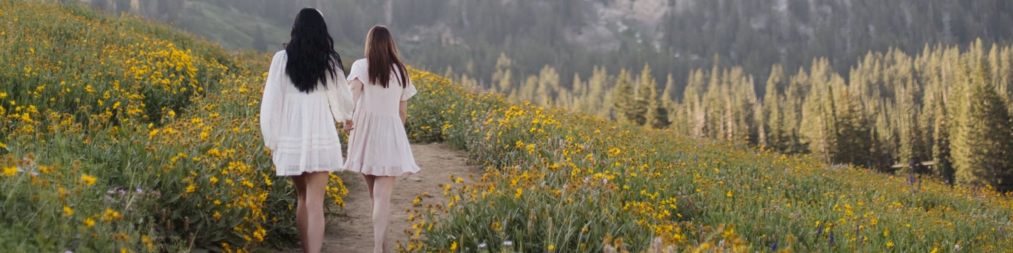 Two women in white dresses walk hand-in-hand down a dirt path through yellow wildflowers, with misty mountains rising behind them.