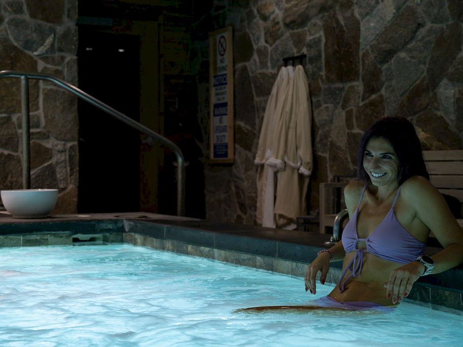 A woman in a purple swimsuit relaxes in an indoor stone-walled pool, smiling while leaning on the edge.