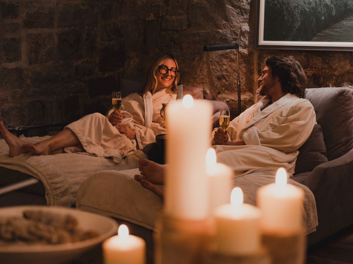 A cozy lounge: two people in robes relax on a couch, toasting with champagne under warm candlelight and soft lights.
