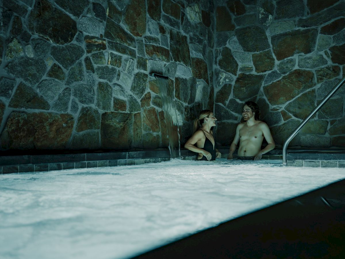 A couple relaxes in a heated indoor pool, under a cascading water feature against a tiled stone wall.