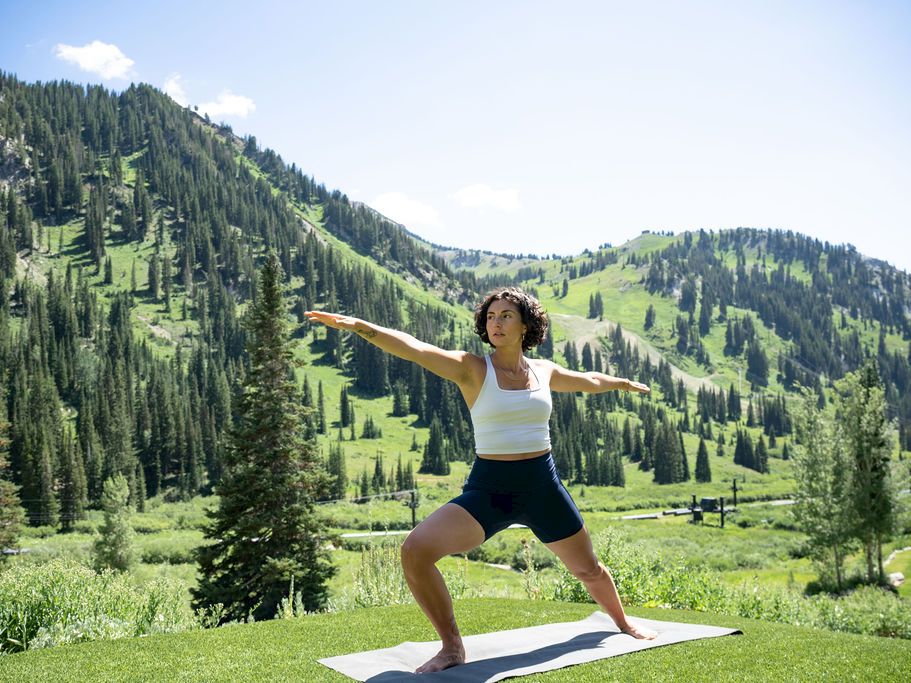 A woman is doing yoga on a mat outdoors, stretching in a lush mountain valley under bright daylight.