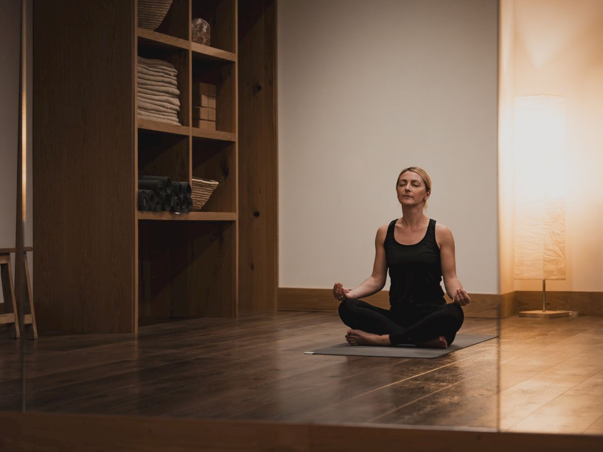 A woman meditates in a calm yoga studio, sitting cross‑legged on a mat with eyes closed.