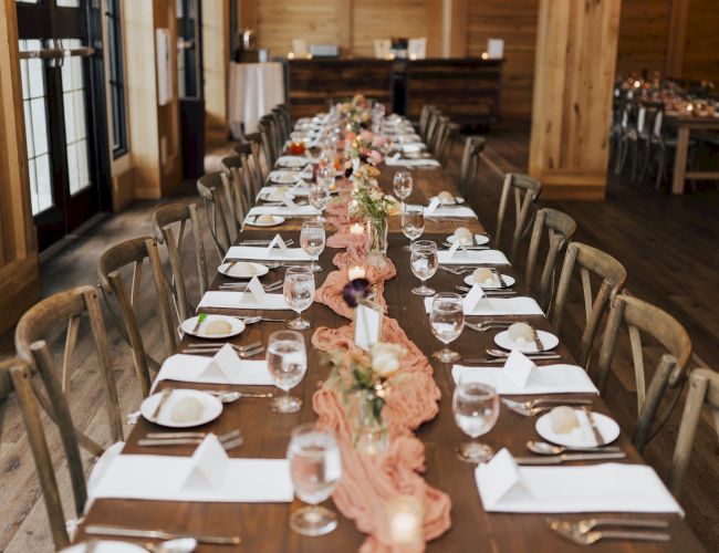A rustic barn dinner setup with long tables, white plates, candles, glasses, and pink floral centerpieces for a celebration.