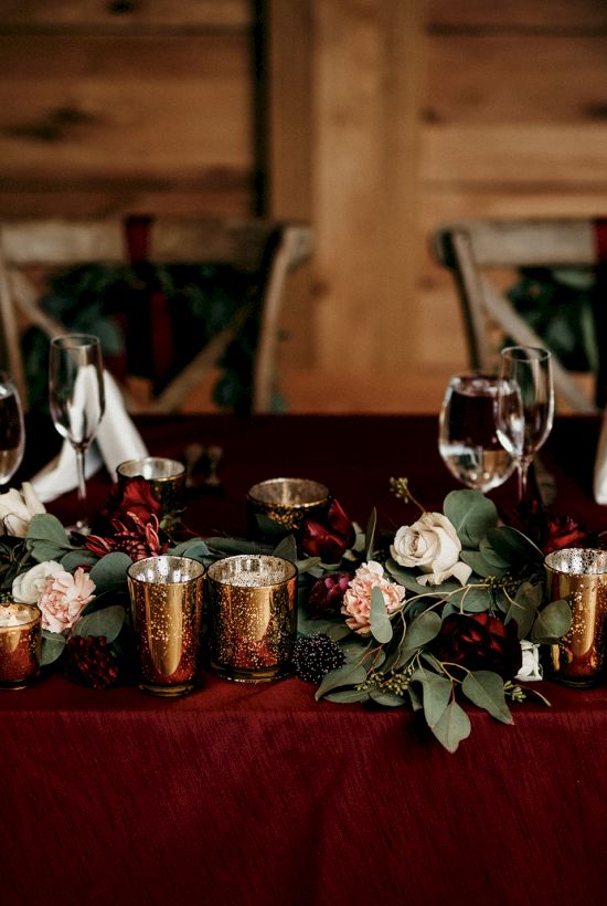 A festive banquet table with copper mugs, wine glasses, candles, and red-and-white flowers in front of a wooden backdrop.
