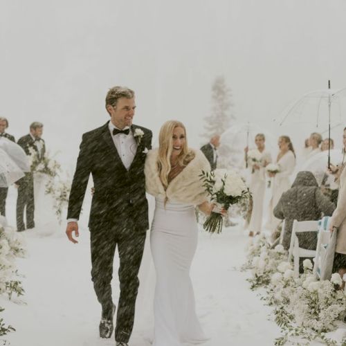 A snowy outdoor wedding procession in white: a groom in a tuxedo and a bride holding flowers walk down the aisle.