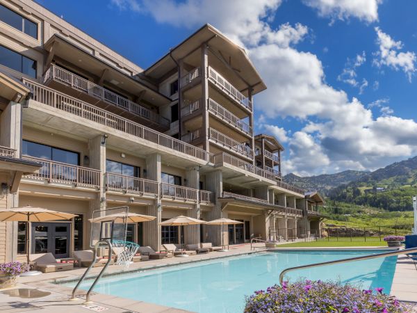 A modern hotel with multiple balconies overlooks a sunny outdoor pool under a blue sky with mountains in the distance.