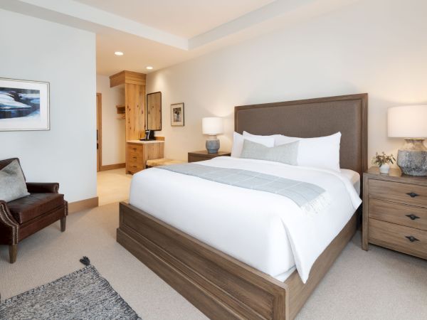 A modern bedroom with a large bed, nightstands and lamps, framed art, a dresser, and a cozy armchair in warm neutral tones.