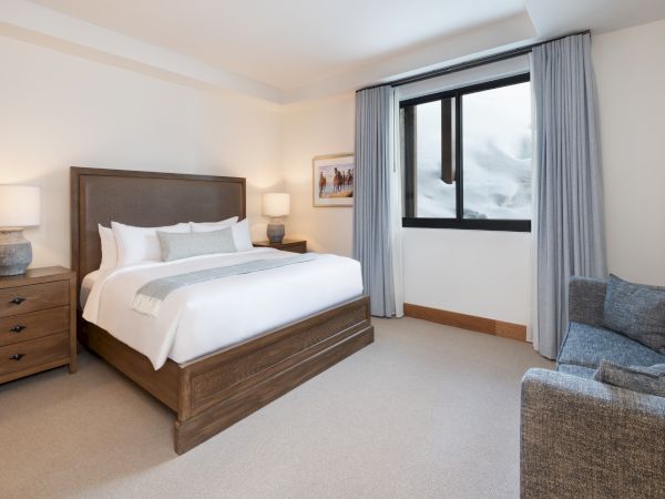A bright modern bedroom with a wooden bed, bedside lamps, gray curtains, and a window showing snowy mountains, with a couch.