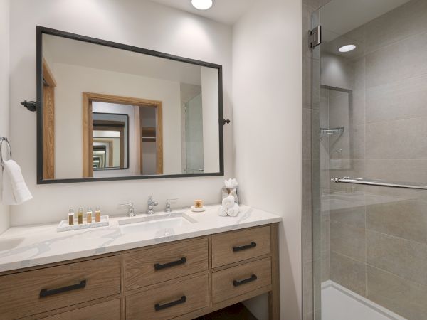 A modern bathroom with a double vanity, large framed mirror, towels, toiletries, and a walk-in glass shower.
