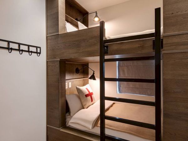 A modern interior with built-in wooden bunk beds, wall lights, a small bed with a red cross pillow, and hanging hooks.