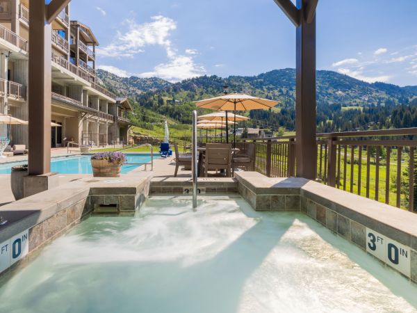 An outdoor hot tub beside a swimming pool, with patio lounge chairs, umbrellas, and mountain views under a clear sky.