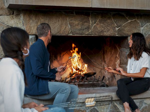 Friends sit by a stone fireplace, roasting marshmallows on sticks and smiling together.