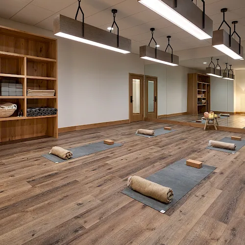 A bright yoga studio with wooden storage shelves, floor mats, folded towels, yoga blocks, and hanging pendant lights.