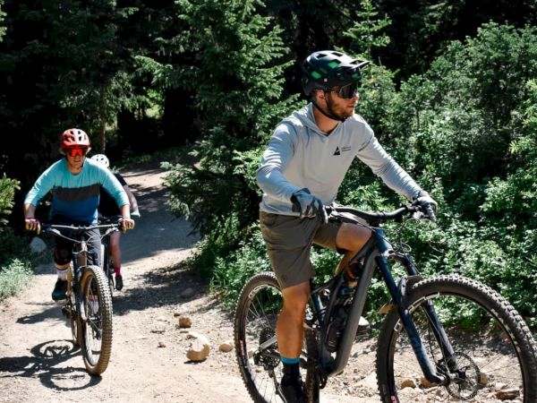 Two mountain bikers ride a sunny dirt path through dense green forest, one in front and one behind.