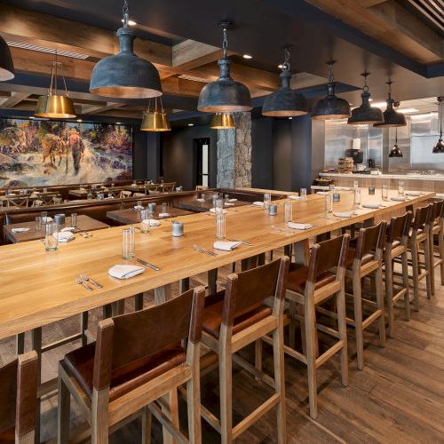 A rustic restaurant dining room with long wooden tables, leather chairs, and hanging industrial pendant lights near an open kitchen.