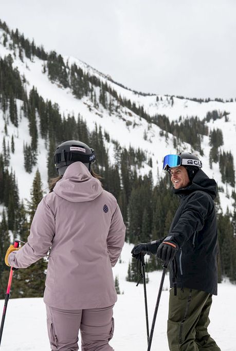 Two skiers in winter gear stand on a snowy mountain slope, chatting and holding ski poles as pine-covered peaks rise behind them.