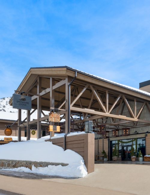 A snowy mountain lodge exterior with the &ldquo;Snowpine Lodge&rdquo; sign and wooden beams under a clear blue sky.