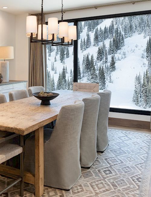 A cozy modern dining room with a large farmhouse table, cushioned chairs, and snowy mountain views through a big window.