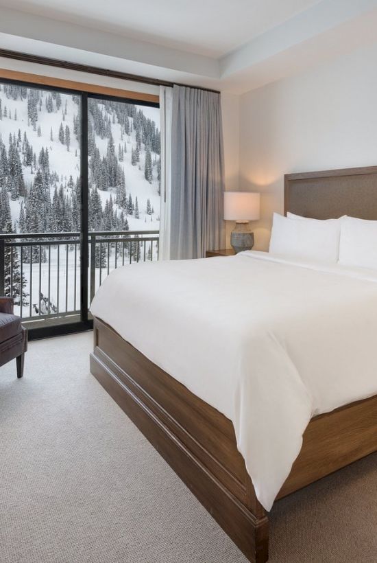 A cozy hotel bedroom with a king bed, bedside tables, and a balcony view of snowy pine trees.