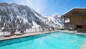 Snowy alpine mountains and pine trees frame an outdoor heated pool beside a stone lodge terrace.