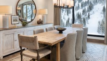 A cozy dining room with a rustic wood table and cushioned chairs, big snowy mountain windows, and warm lamps.