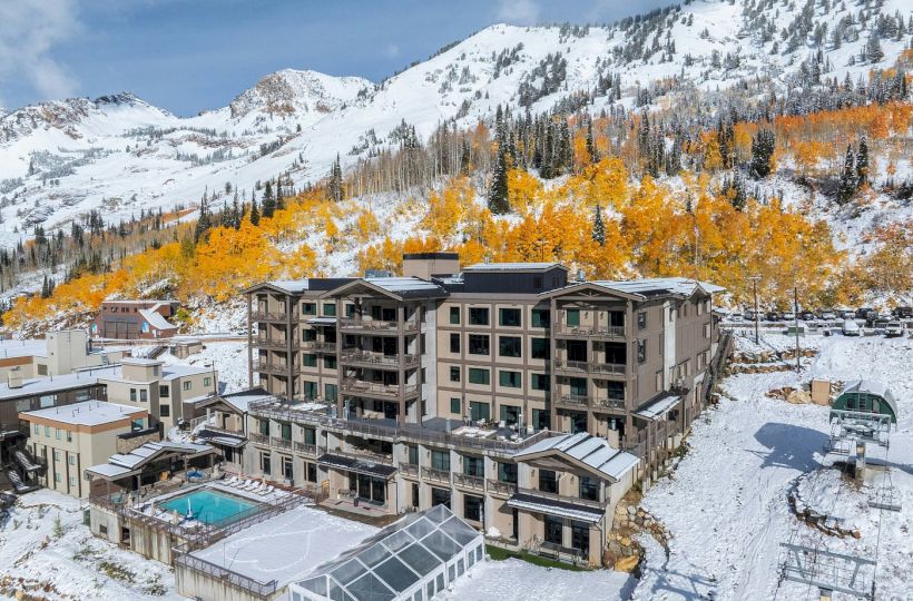 A mountain resort hotel with an outdoor pool sits amid snow-covered peaks and orange trees under a cloudy sky.