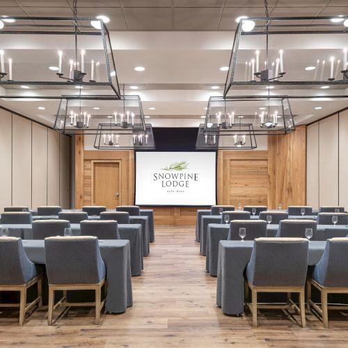 An elegant lodge conference room with rows of gray chairs, hanging lantern-style chandeliers, and a &ldquo;Snowpine Lodge&rdquo; stage screen.