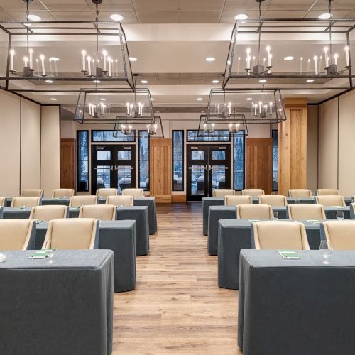 A modern meeting room with rows of gray tables, beige chairs, and hanging candle-style chandeliers.
