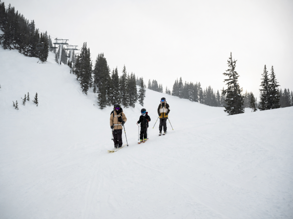 Three skiers hike downhill on a snowy mountain slope past pine trees, with a chairlift in the background.