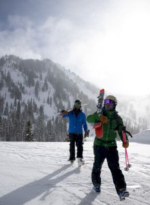 Two snowboarders in colorful gear stand on a snowy piste with skis and mountains behind them, snow falling and clouds clearing.