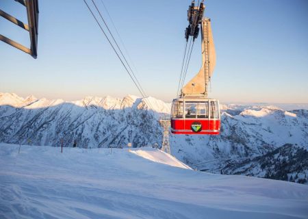 A red gondola cable car swings over snowy ski slopes with towering mountains in the background at sunset.