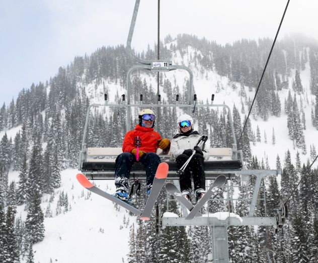 Two skiers ride a chairlift above snowy pine forests, wearing colorful helmets and goggles, in a foggy winter mountain landscape.