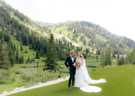 A bride and groom pose outdoors on a green hillside with fir forests and mountains in the background.