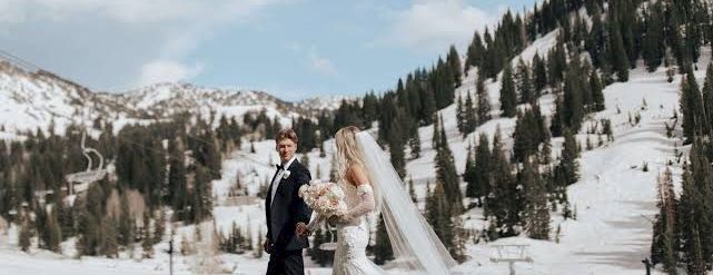 A bride and groom in wedding attire pose on a grassy field with snowy alpine mountains and pine trees in the background.