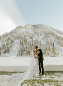 A bride and groom kiss outdoors in snowy Alps, their wedding portrait set against pine trees and mountains.