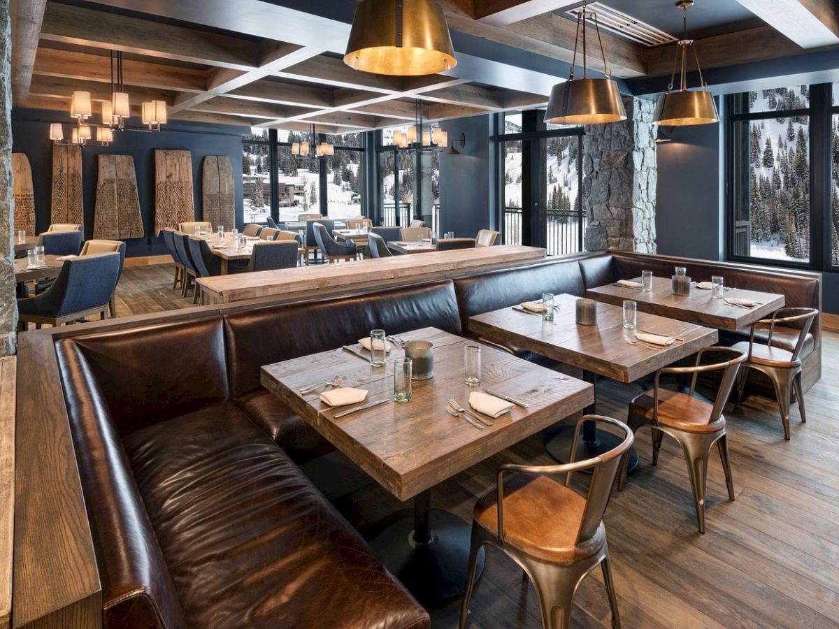 A modern ski-lodge dining room with wooden tables, leather booths, pendant lights, and large windows showing snowy pine trees.