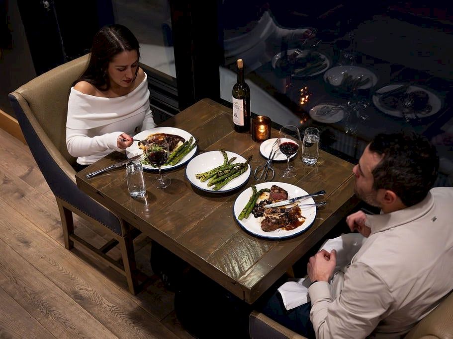 Two people dine at a table with steak and asparagus plates, wine bottles, and candlelight.