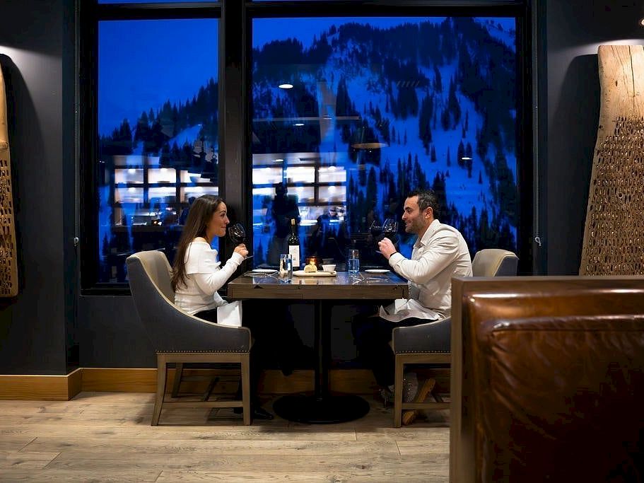 Two people in a restaurant booth toast with drinks across a table, while big windows show a blue mountain night outside.
