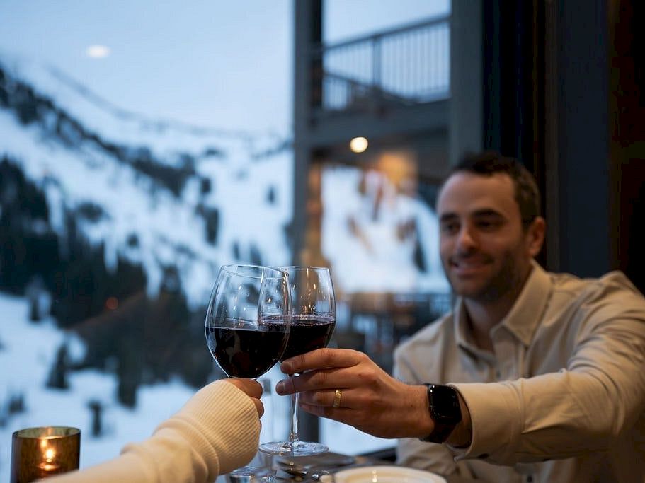 A man cheers with a companion, clinking red wine glasses at a cozy restaurant with snowy street views behind them.