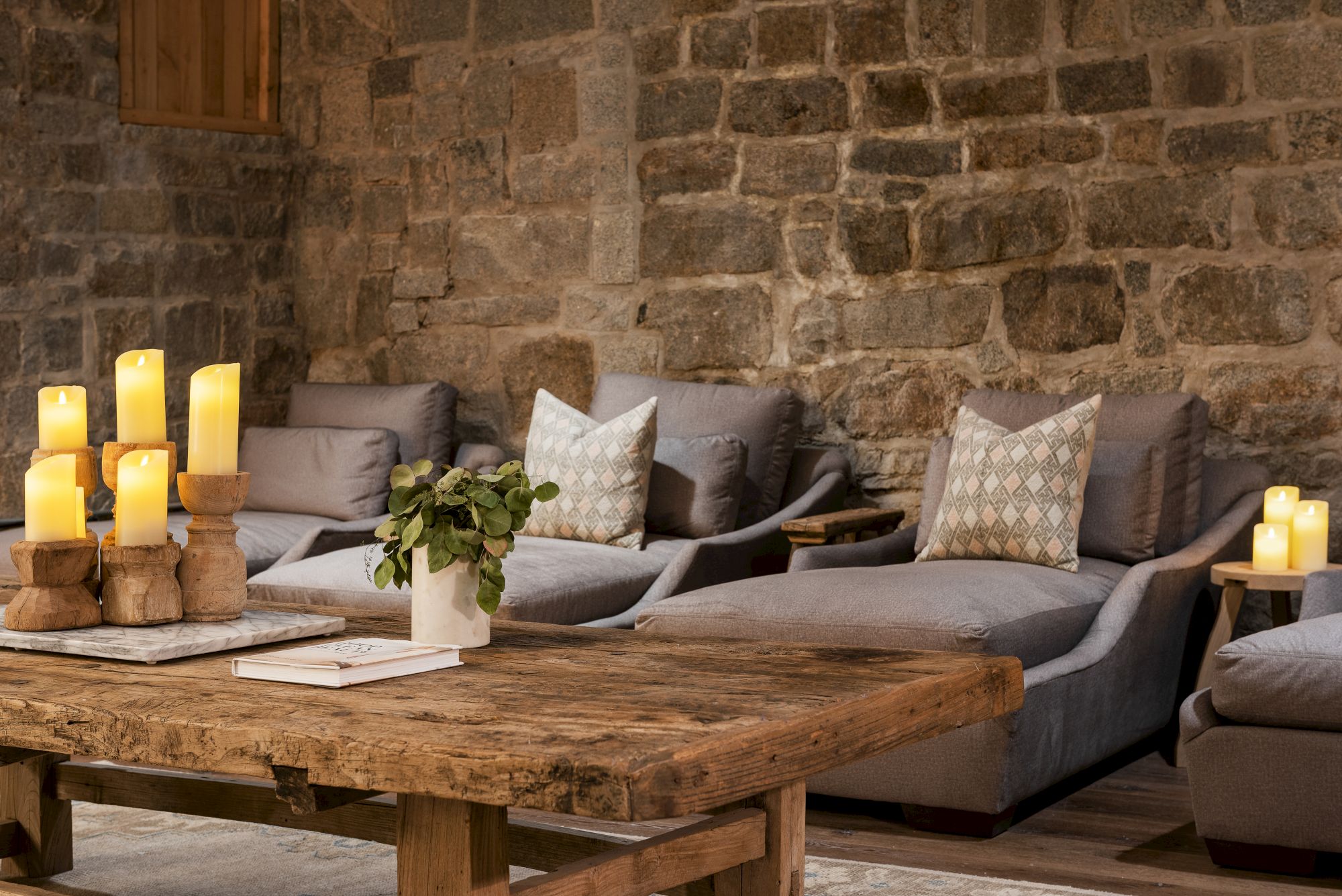 A cozy stone-walled lounge with a sectional couch, pillows, lit candles, and a wooden coffee table for warm ambiance.