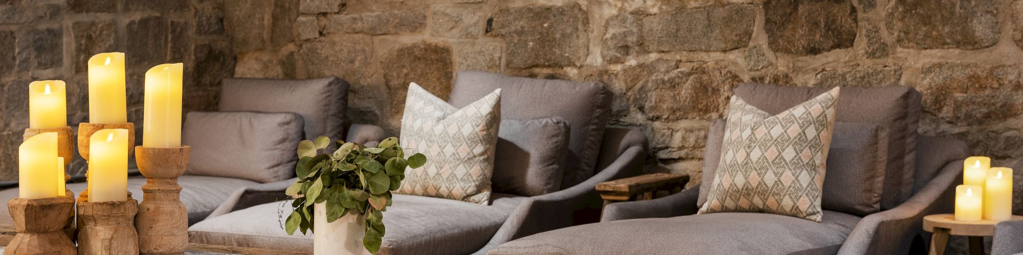A cozy stone-walled lounge with a sectional couch, pillows, lit candles, and a wooden coffee table for warm ambiance.